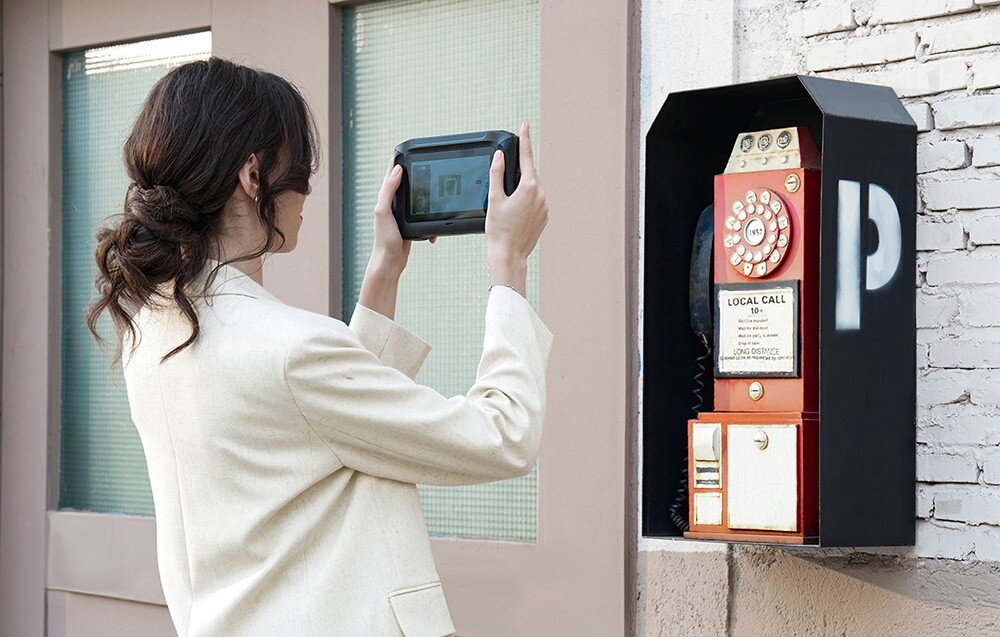 Osoba trzyma urządzenie przypominające skaner 3D przed czerwonym, zabytkowym telefonem publicznym z tarczą obrotową i napisem \'LOCAL CALL\'.