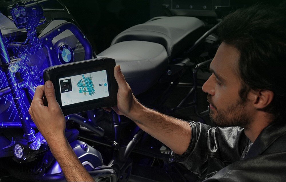 Osoba trzyma urządzenie z wyświetlaczem, na którym widoczny jest model 3D motocykla. W tle znajduje się motocykl z logo BMW.
