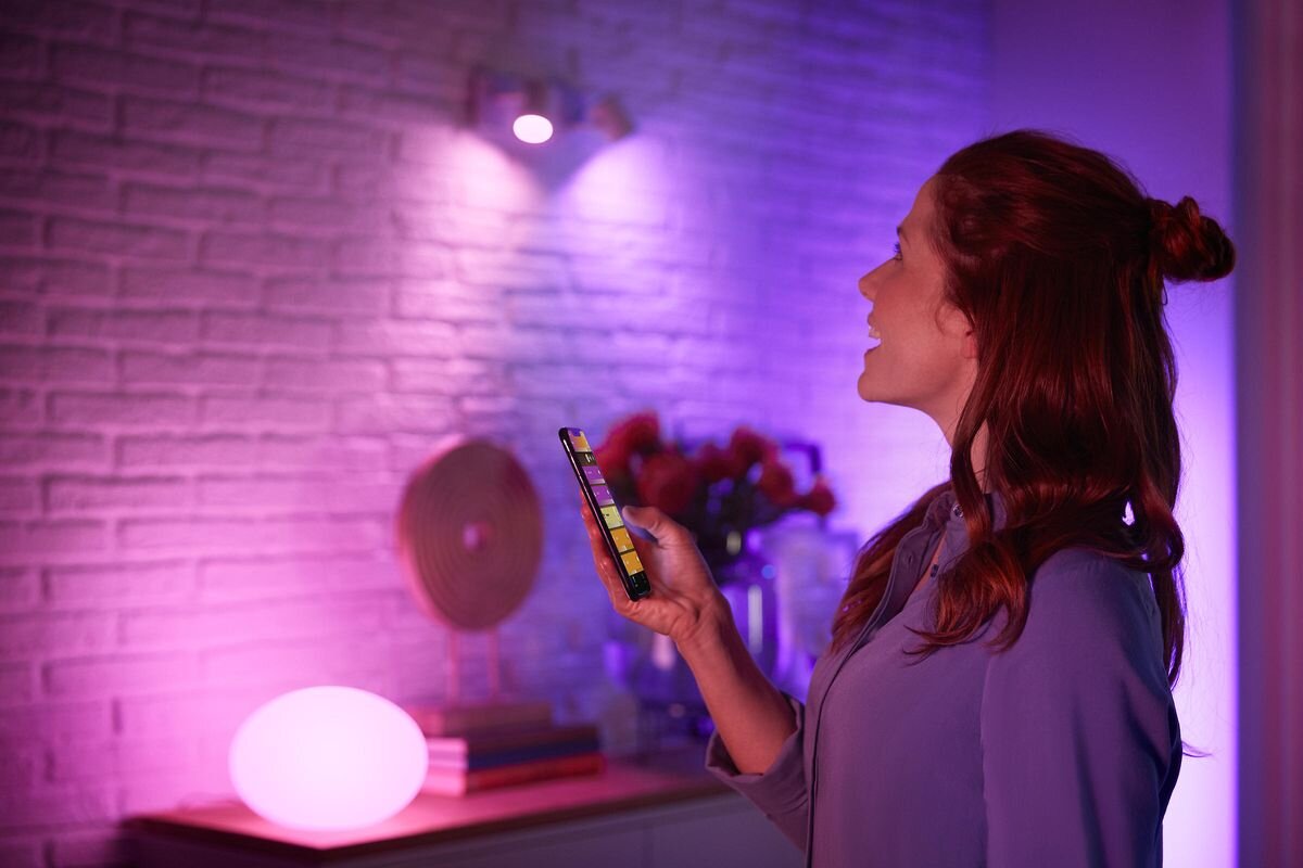 Lampa biurkowa PHILIPS HUE Flourish (Bluetooth, ZigBee), głosowe, HomeKit, Alexa, Google, Kobieta sterująca telefonem kolorowym oświetleniem Philips Hue w domu.