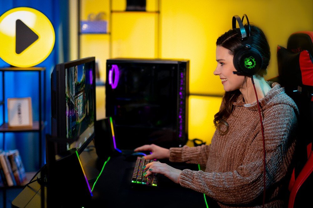 Komputer MAD DOG Powered By Asus GT302-A01WR32WH Uśmiechnięta kobieta w słuchawkach z logo świecącym na zielono siedzi przy biurku i trzyma dłonie na klawiaturze z kolorowym podświetleniem