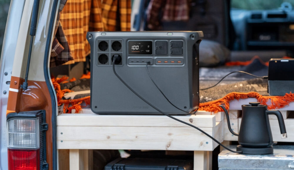 Stacja zasilania DJI Power 2000 2048 Wh Bank energii, naładowanie, smartfon, urządzenia, pojemność, w tle otwarty bagażnik auta, stacja zasila czajnik stojący obok na stole