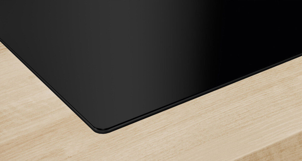 PŁYTA SIEMENS EE611BPB5E Na zdjęciu pokazano zbliżenie narożnika czarnej, szklanej płyty grzewczej osadzonej w jasnym, drewnianym blacie. Gładka, równo wkomponowana krawędź podkreśla precyzyjne wykończenie i elegancki wygląd zabudowy