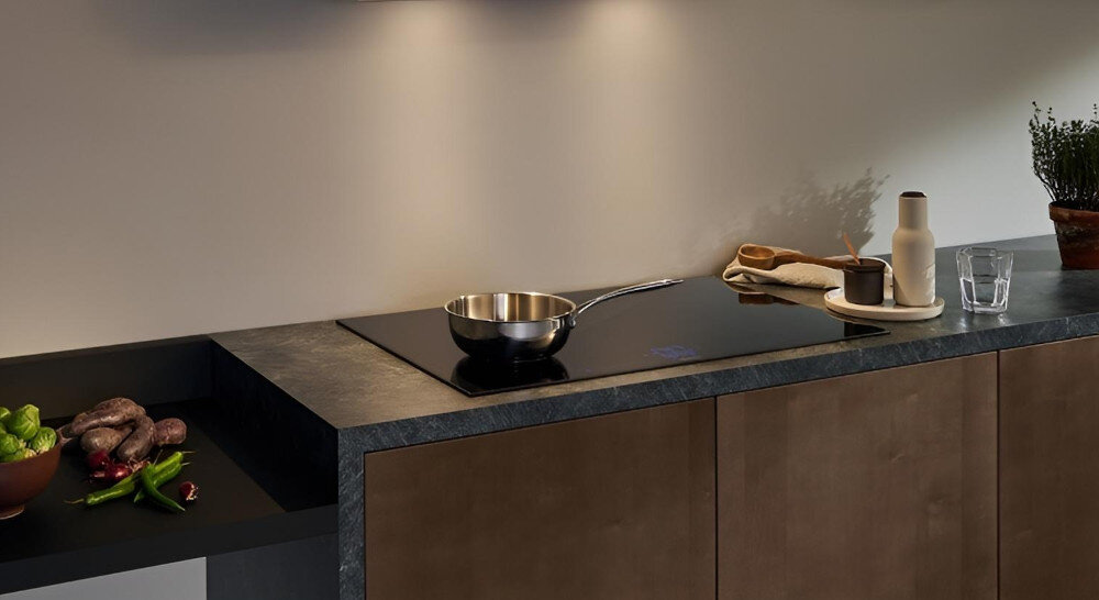 PŁYTA SIEMENS EE611BPB5E Na zdjęciu widać kuchnię z czarnym blatem, w który wkomponowana jest czarna płyta indukcyjna z pojedynczym rondlem ustawionym na środku. Delikatne oświetlenie podszafkowe oraz odłożone na blacie akcesoria i zioła tworzą ciepłą, minimalistyczną aranżację blokada ochrona dzieci bezpieczeństwo