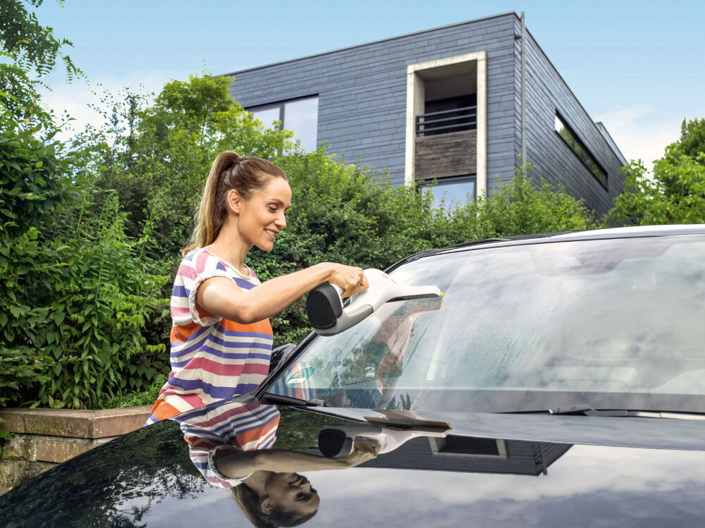 Osoba używa myjki do okien na przedniej szybie samochodu, w tle widać nowoczesny budynek i zieleń.