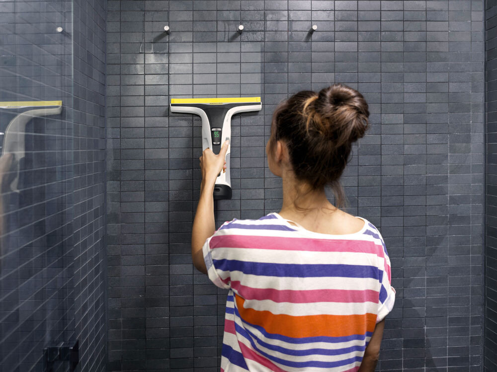 Osoba używa myjki do okien na ciemnej, kafelkowej ścianie. Myjka ma żółty element na górze i jest trzymana w jednej ręce.