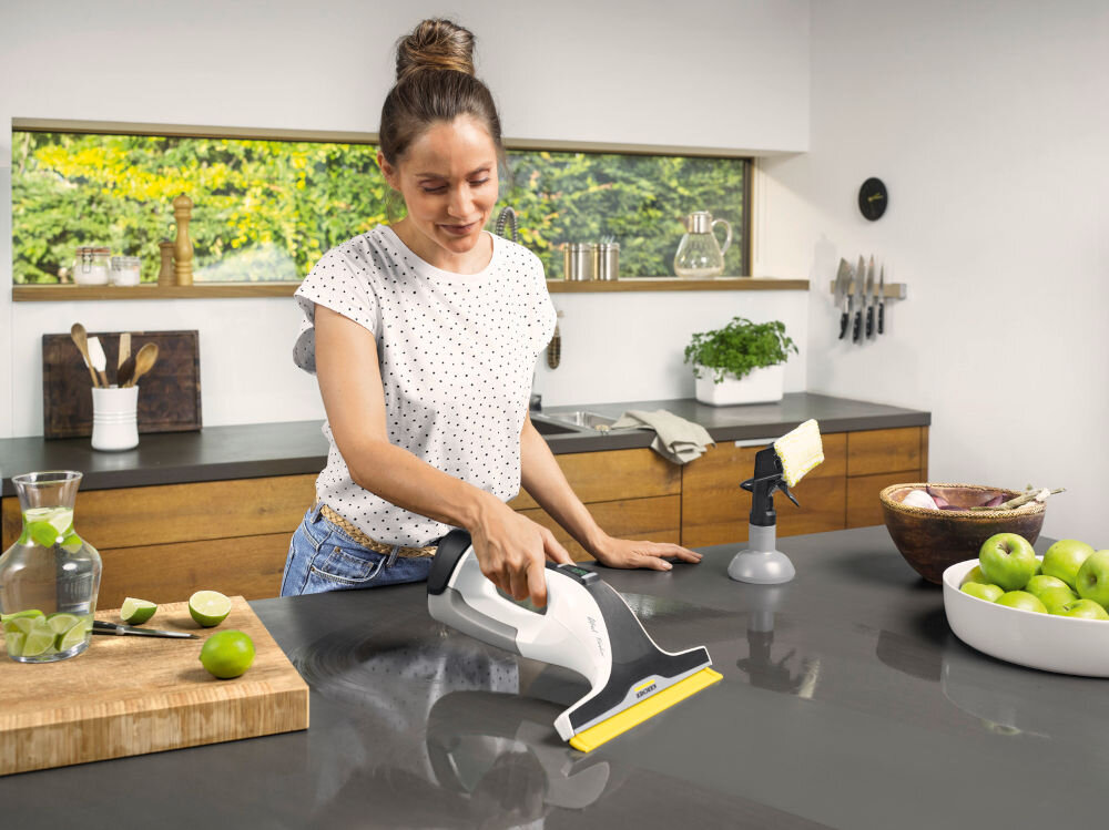 Osoba używa myjki do okien na blacie kuchennym. W tle widać drewniane szafki, misę z zielonymi jabłkami i deskę z pokrojonymi limonkami.