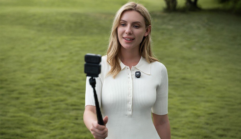 Kamera sportowa DJI Osmo Nano Kobieta stojąca na trawie, trzymająca kamerę na wysięgniku, druga przypięta do koszulki na piersi, wbudowane mikrofony, dźwięk stereo, system OsmoAudio