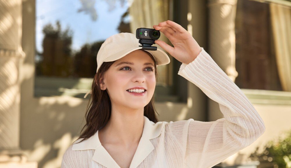 Kamera sportowa DJI Osmo Nano Kobieta w jasnej czapce z daszkiem z przypiętą na froncie małą kamerą, uśmiechająca się i unosząca rękę, konstrukcja, waga, magnetyczne mocowanie