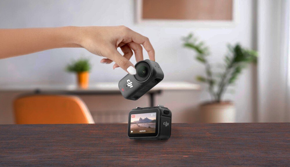 Kamera sportowa DJI Osmo Nano Ręka kobiety trzymająca niewielką kamerę nad drugą stojącą na drewnianym blacie, w tle wnętrze mieszkania, stacja dokująca, ekran OLED, obracanie