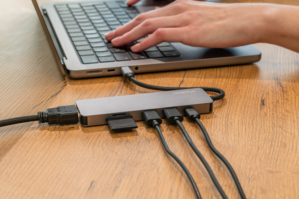Hub USB XQUANTUM UH210 6w1 hub podpięty do komputera przez port USB-C obok 4 wtyczki USB-A ułożone obok portów adaptera