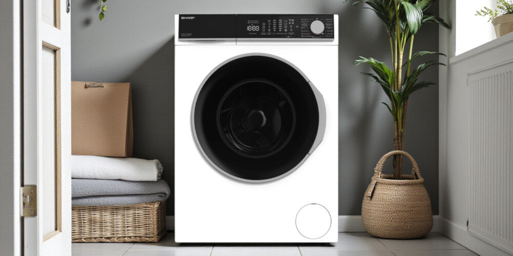 PRALKA SHARP ES-NFL714AWNNA-PL Na zdjęciu znajduje się biała pralka marki SHARP ustawiona na środku jasnej pralni. Wokół niej widać kosz z ręcznikami, roślinę w wiklinowej donicy oraz drzwi i grzejnik, co nadaje wnętrzu domowy charakter inteligentne pranie eco track eco logic allergy smart funkcja pary