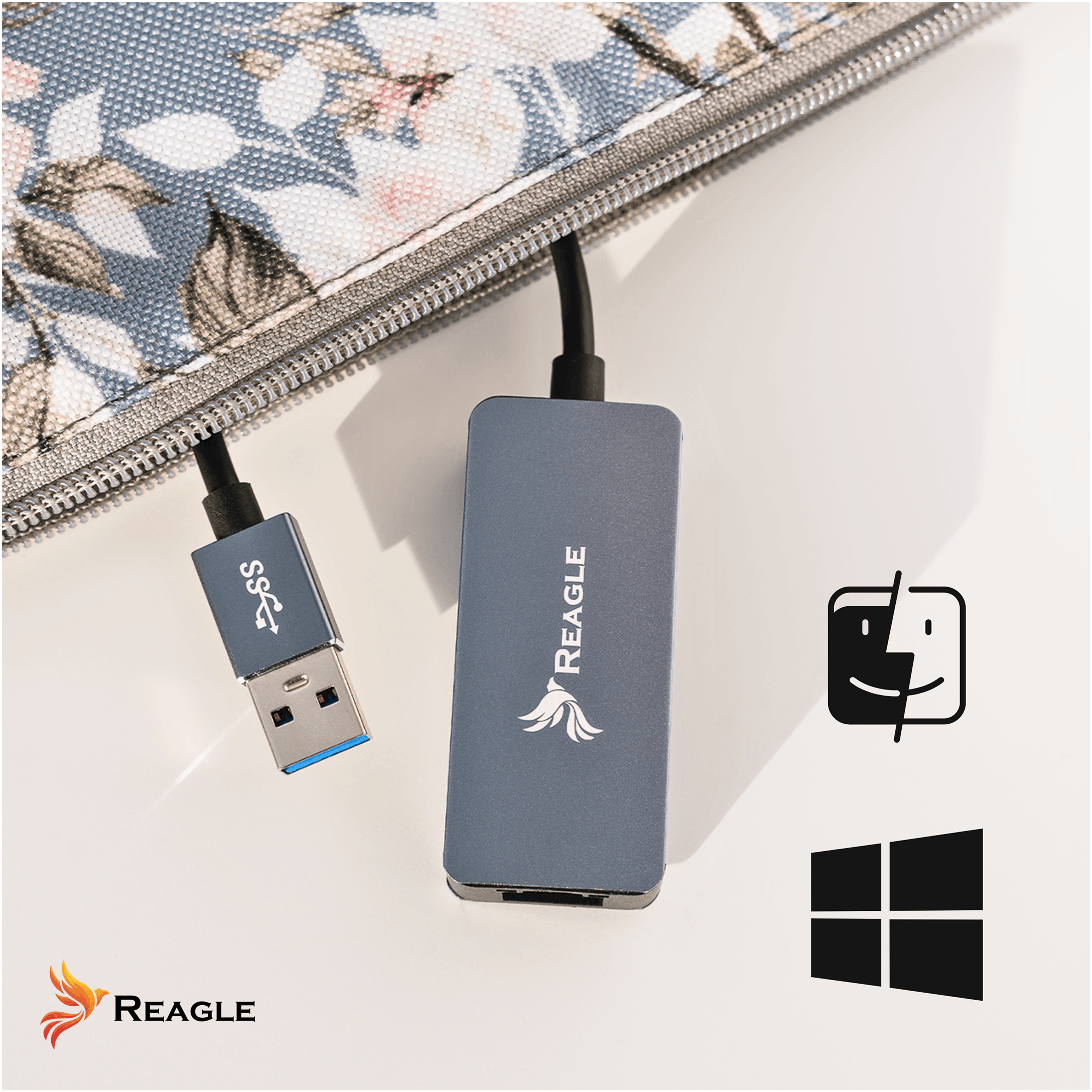 Na obrazie widoczny jest adapter USB z logo REAGLE, obok którego znajdują się ikony systemów operacyjnych. W tle znajduje się materiał z kwiatowym wzorem.