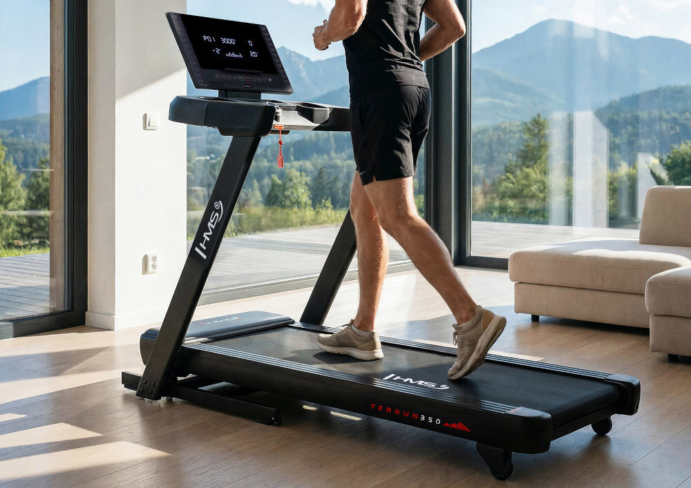Bieżnia elektryczna HMS TERRUN350 1305W 22 km-h dom pokój osoba korzysta z bieżni w tle duże okno kanapa dwukierunkowy trening w domu ćwiczenia dbanie o kondycję