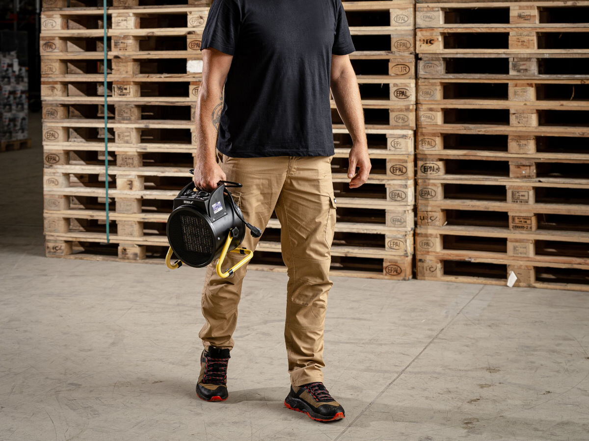 Nagrzewnica elektryczna DEDRA DED9931C, uchwyt, masa, ergonomia, mobilność, Mężczyzna niesie nagrzewnicę DED9931C w magazynie, kratkowana siatka, żółty stelaż, czarna obudowa.