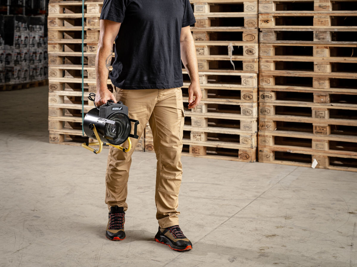 Nagrzewnica elektryczna DEDRA DED9930B, lekkość, uchwyt, cicho, czysto, Mężczyzna w beżowych spodniach niesie nagrzewnicę Dedra DED9930B po magazynie z paletami.