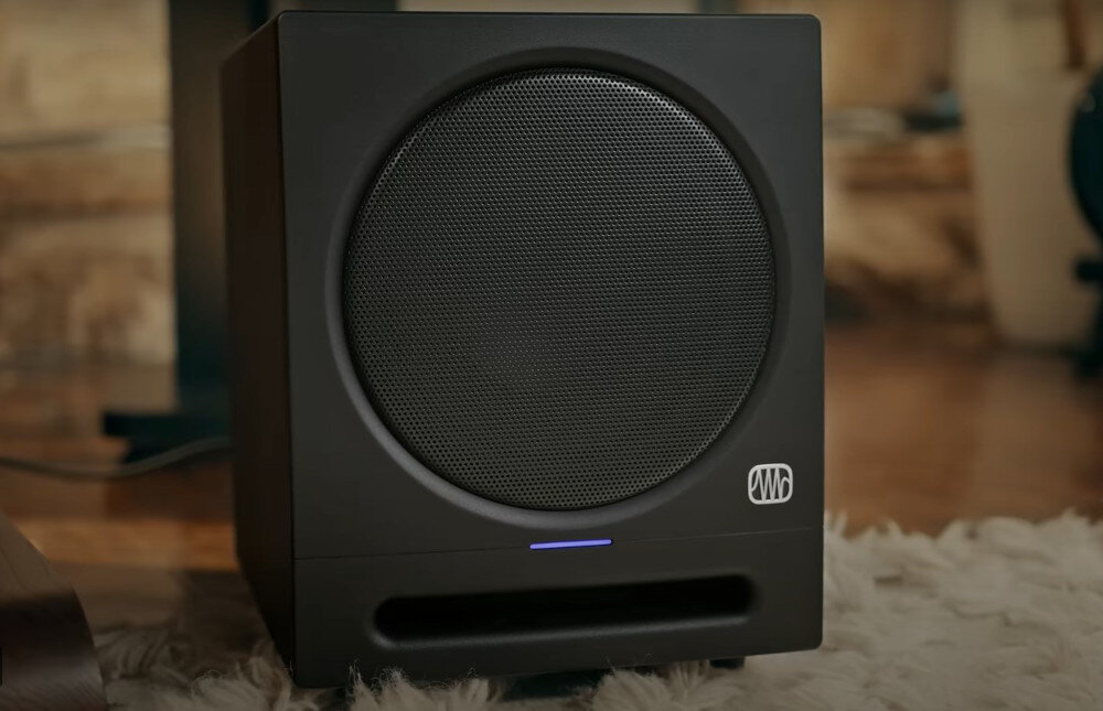 Czarny subwoofer z metalową siatką na przedniej części i niebieskim wskaźnikiem LED. Na obudowie widoczne jest logo producenta.