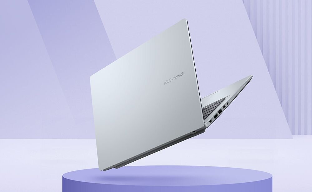 Srebrny laptop z widocznym logo \'ASUS Vivobook\' na pokrywie, umieszczony na fioletowym tle. Laptop jest lekko otwarty, ukazując porty po jednej stronie.