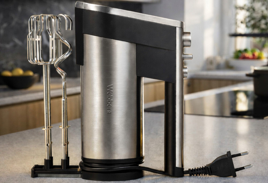 Na blacie kuchennym stoi mikser o metalicznym wykończeniu z widocznym napisem \'WEBBER\'. Obok miksera znajdują się dwa mieszadła oraz przewód zasilający z wtyczką.