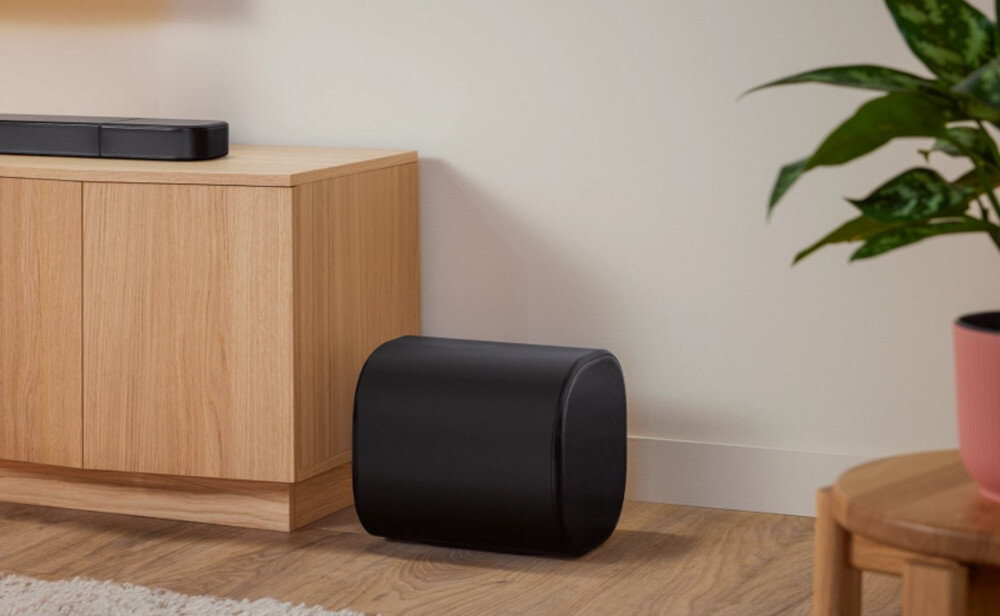 Na zdjęciu widać czarny soundbar i subwoofer ustawione obok drewnianej szafki na tle jasnej ściany. Obok znajduje się roślina w doniczce na drewnianym stoliku.