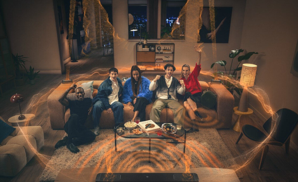 Grupa osób siedzi na kanapie w salonie, przed nimi na stoliku znajdują się przekąski. W pomieszczeniu widoczny jest efekt wizualny przypominający fale dźwiękowe.