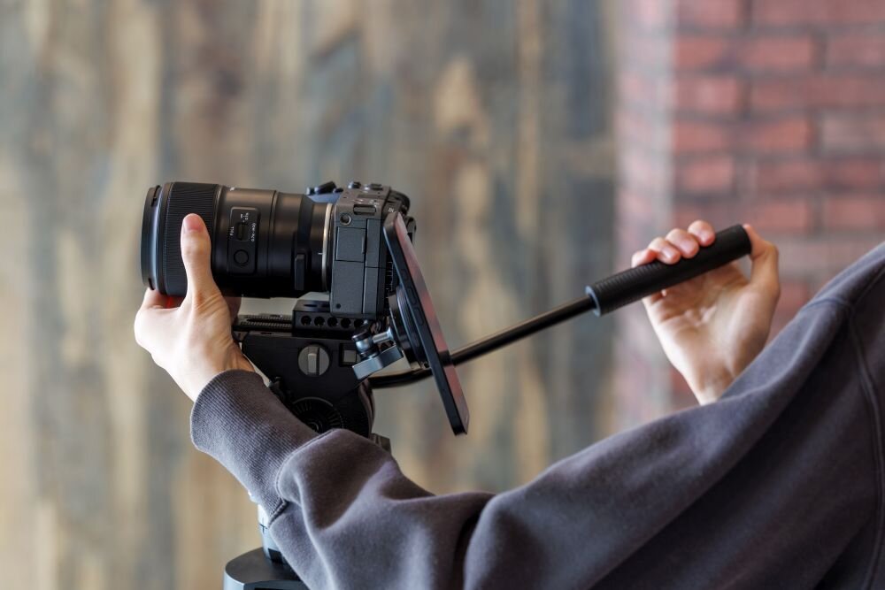 Osoba trzyma aparat fotograficzny zamontowany na statywie z uchwytem. W tle widoczna jest rozmyta ściana z cegieł i drewna.