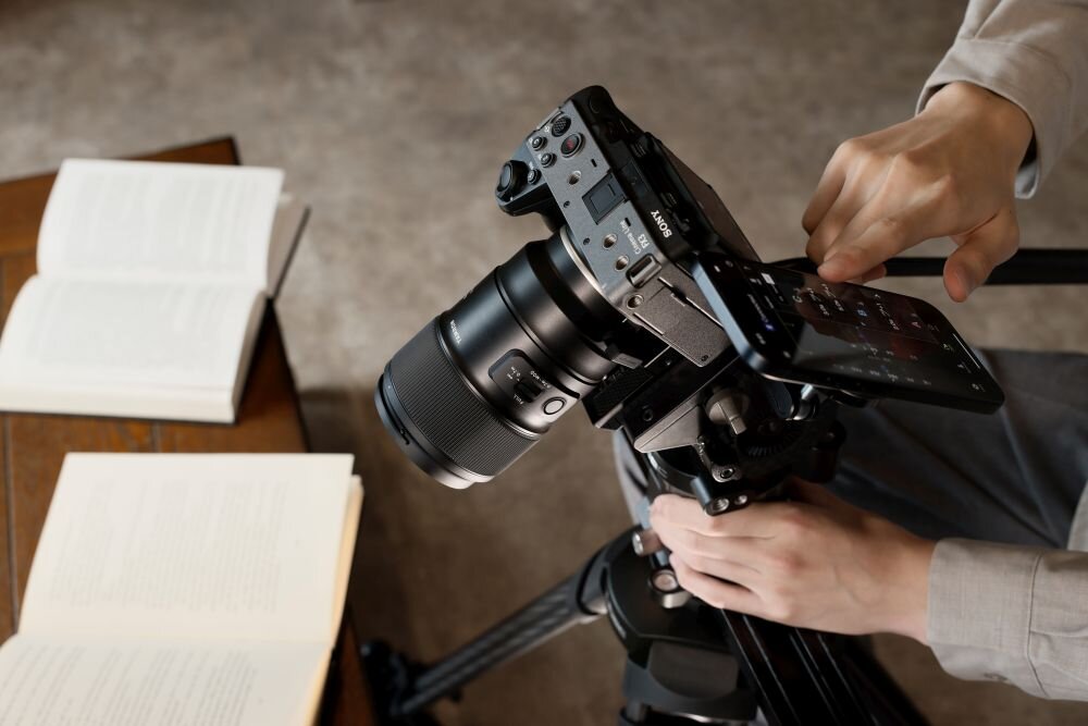 Osoba obsługuje aparat fotograficzny zamontowany na statywie, obok leżą otwarte książki. Na aparacie widoczne jest logo \'SONY\'.