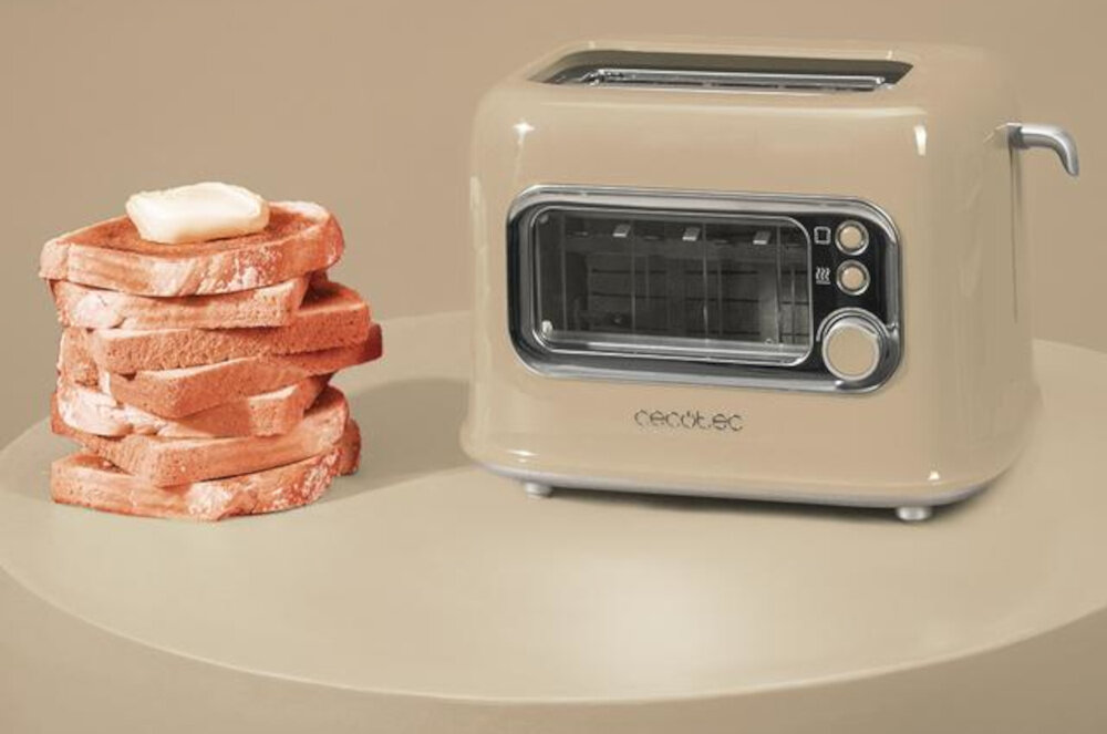Beżowy toster z przezroczystym panelem i trzema przyciskami znajduje się obok stosu tostów z masłem. Na tosterze widoczne jest logo \'cecotec\'.