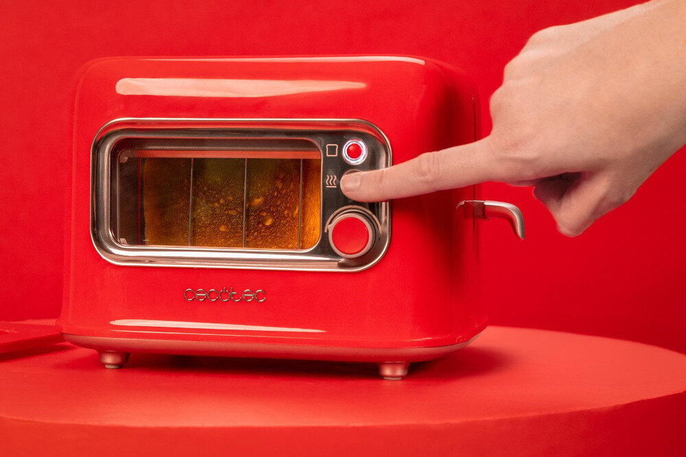 Czerwony toster z przezroczystym okienkiem, przez które widać opiekany chleb. Na obudowie widoczne jest logo \'cecotec\', a ręka naciska przycisk na panelu sterowania.