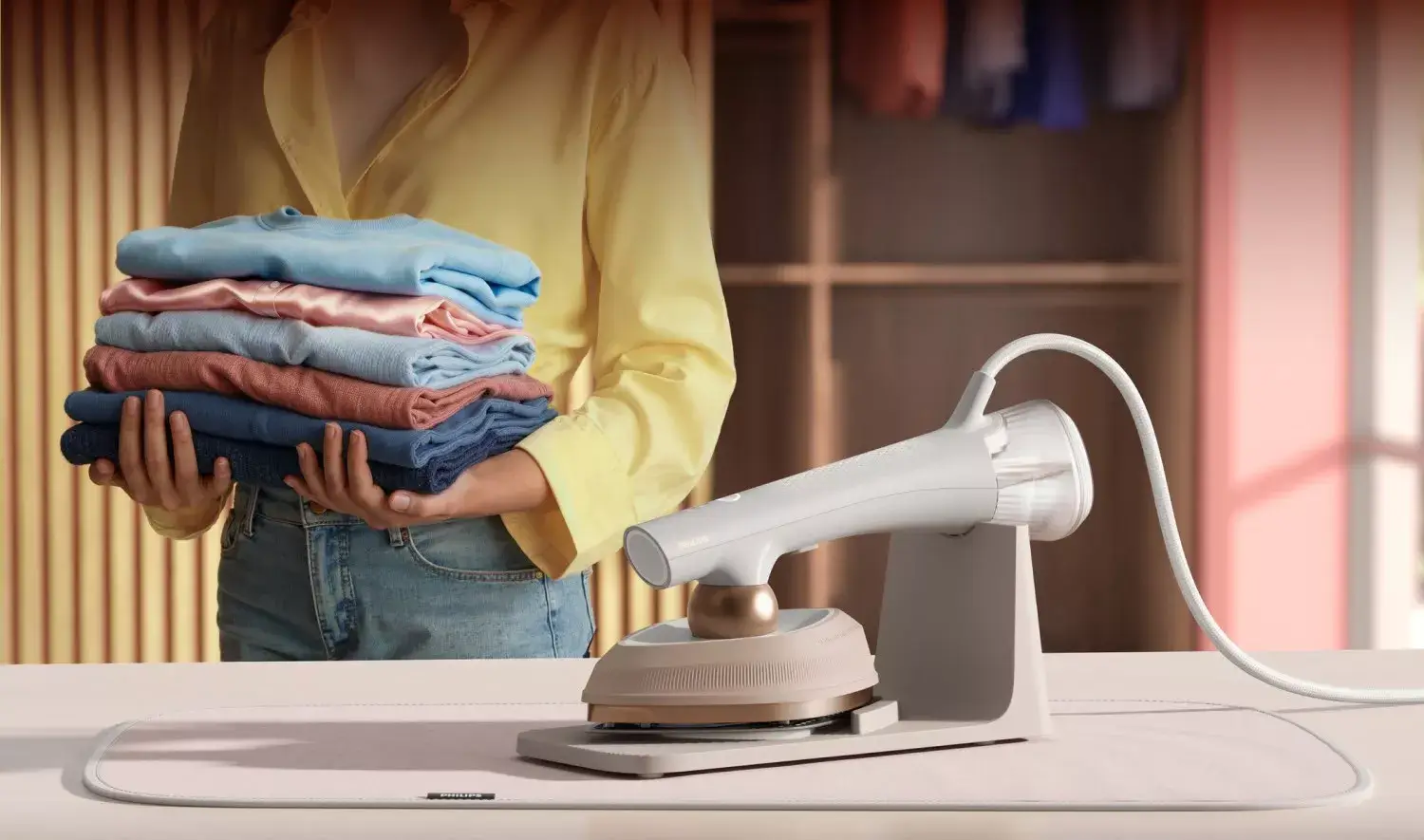 Osoba trzyma stos złożonych ubrań w różnych kolorach. Obok znajduje się parownica do ubrań na podstawce.