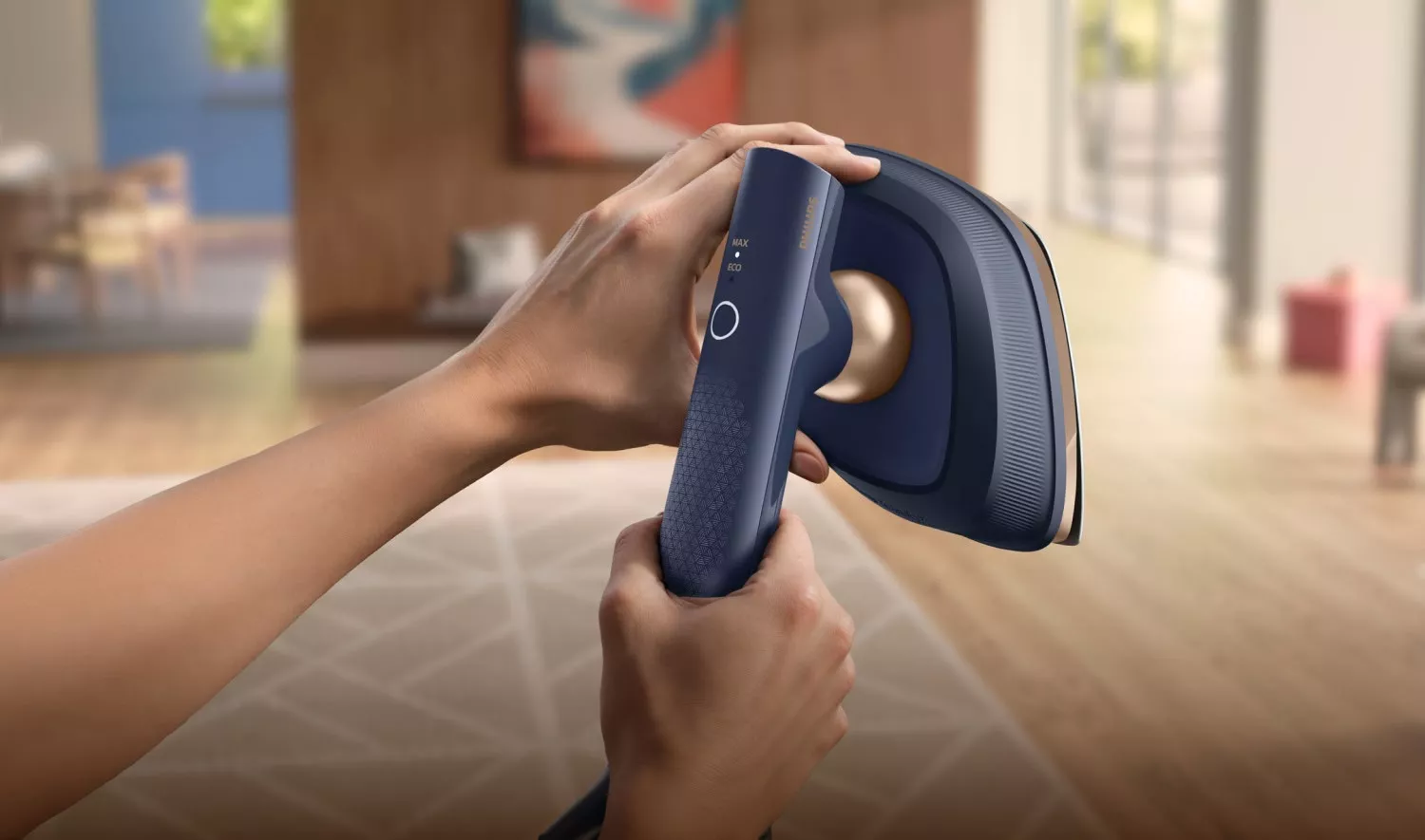 Osoba trzyma w rękach niebieską parownicę do ubrań z widocznym logo \'PHILIPS\'. W tle znajduje się wnętrze pokoju z rozmytymi elementami wystroju.