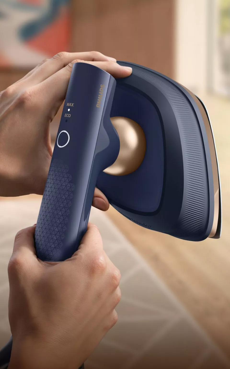 Osoba trzyma w rękach niebieską parownicę do ubrań z widocznym logo \'Philips\'. Na urządzeniu znajdują się oznaczenia \'MAX\', \'ECO\' oraz okrągły przycisk.