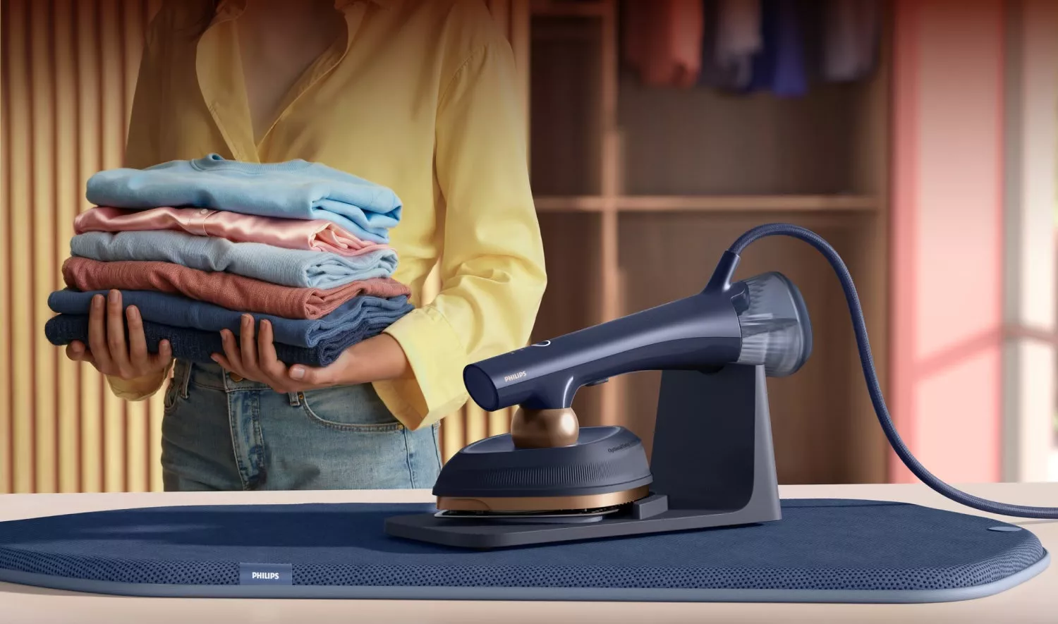 Na obrazie widać osobę trzymającą stos złożonych ubrań oraz urządzenie do prasowania z logo PHILIPS na desce do prasowania. W tle znajduje się szafa z ubraniami.