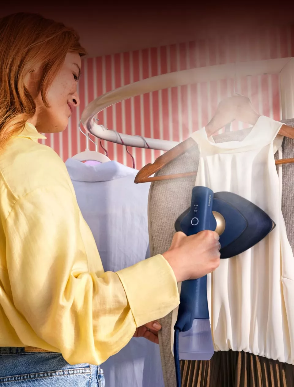 Osoba używa parownicy do ubrań na białej bluzce wiszącej na wieszaku. Parownica jest koloru niebieskiego z widocznym logo PHILIPS.