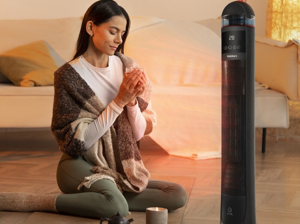 Na obrazie widać osobę siedzącą na podłodze w przytulnym wnętrzu, trzymającą kubek. Obok znajduje się czarny grzejnik ceramiczny z wyświetlaczem pokazującym temperaturę 28 i logo Webber.