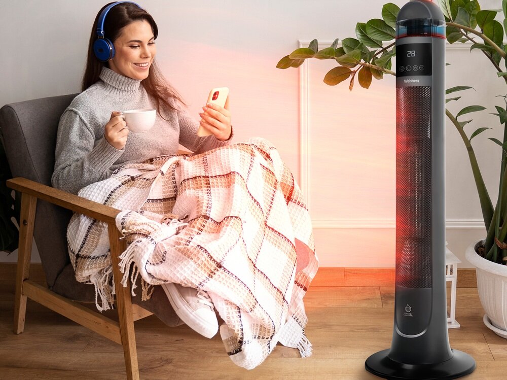 Osoba siedzi na fotelu z kubkiem i telefonem, przykryta kocem. Obok stoi grzejnik ceramiczny z wyświetlaczem pokazującym \'28\' i logo \'Webber\'.