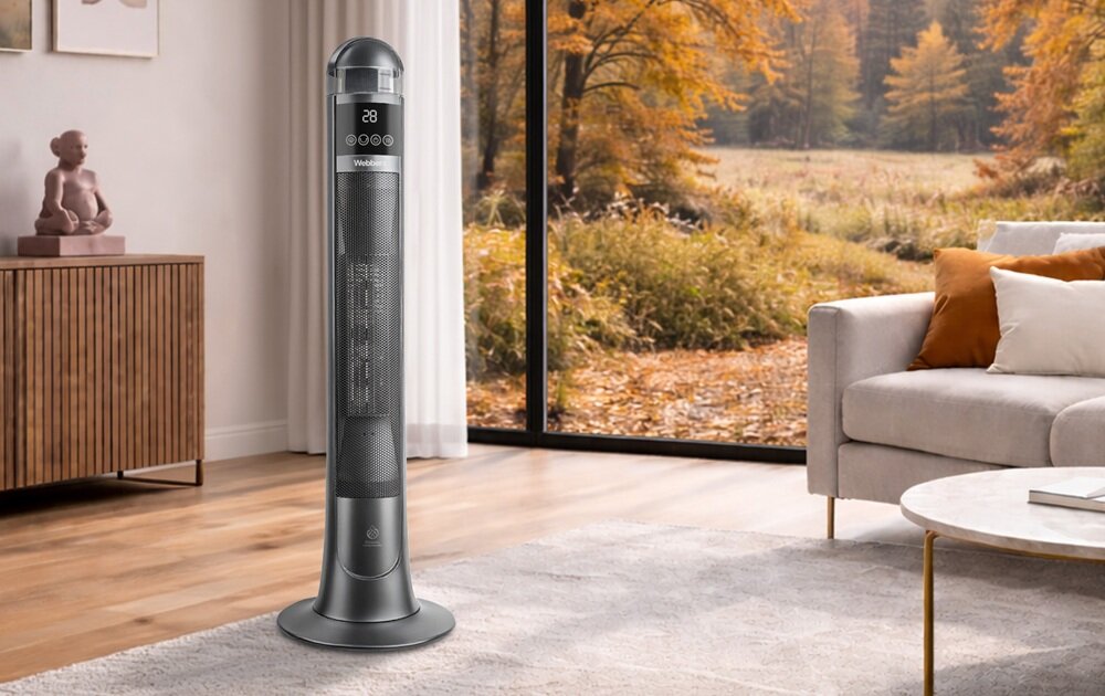 W salonie z dużym oknem stoi wysoki, szary grzejnik ceramiczny z wyświetlaczem pokazującym \'28\' i kilkoma przyciskami. W tle widać sofę, stolik i jesienny krajobraz za oknem.