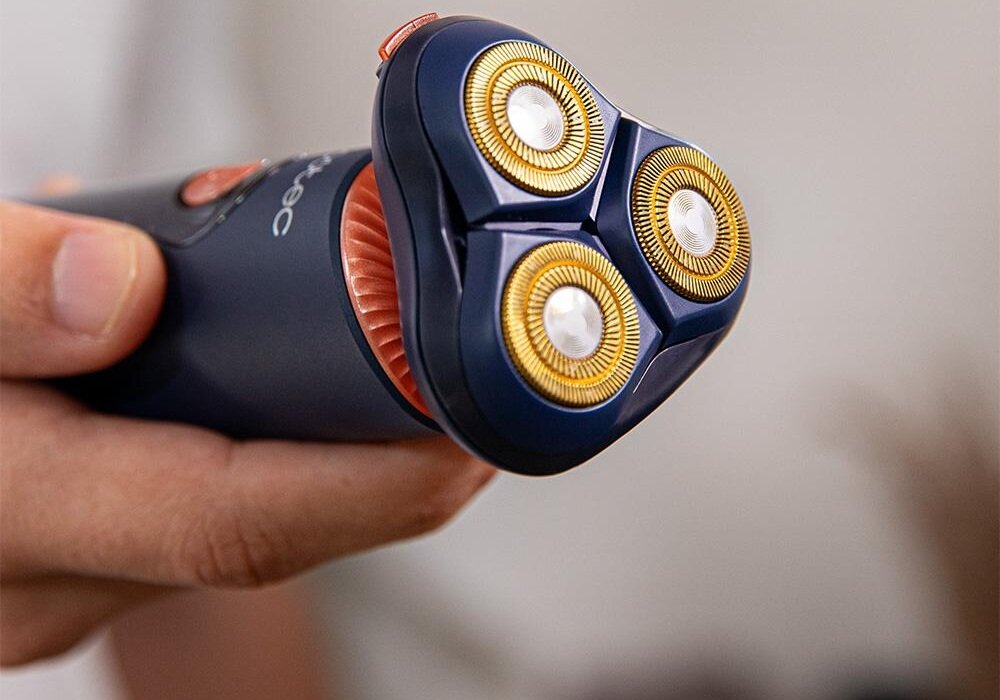 Osoba trzyma elektryczną golarkę z trzema okrągłymi głowicami tnącymi w kolorze złotym. Na uchwycie widoczne jest logo \'cecotec\'.