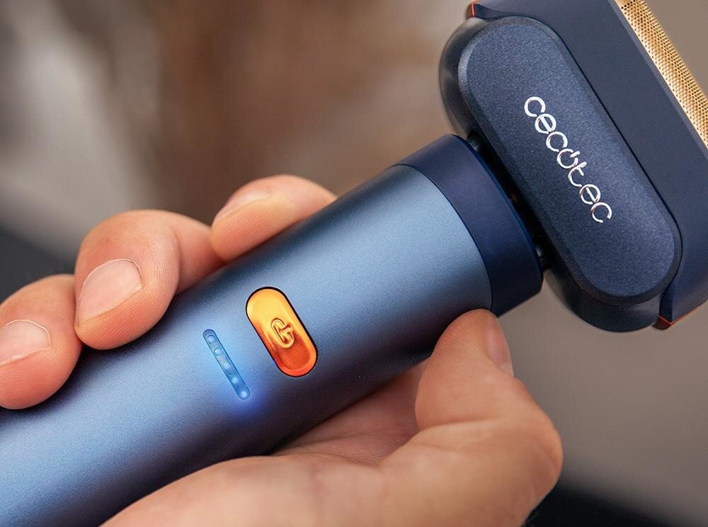Osoba trzyma golarkę w kolorze niebieskim z pomarańczowym przyciskiem i logo \'cecotec\' na głowicy. Na uchwycie widoczne są niebieskie diody LED.