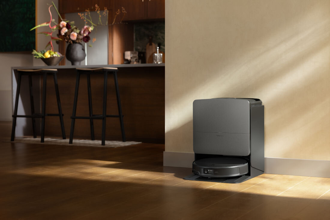 Czarny robot sprzątający znajduje się w stacji dokującej przy ścianie w nowoczesnym wnętrzu. W tle widoczny jest fragment kuchni z wyspą i dwoma krzesłami barowymi.