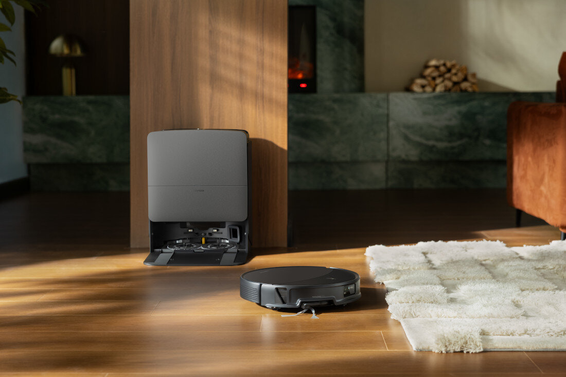 Na drewnianej podłodze znajduje się czarny robot sprzątający obok stacji dokującej. W tle widoczny jest fragment salonu z dywanem i kominkiem.