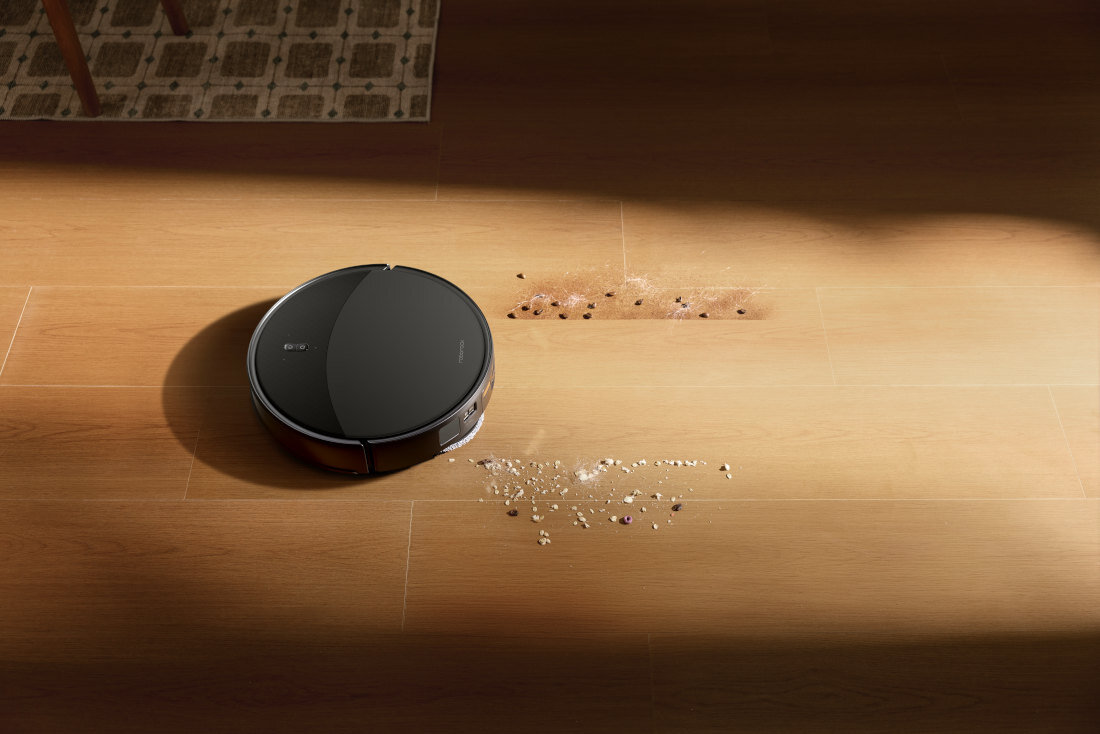 Czarny robot sprzątający na drewnianej podłodze, obok rozsypanych drobinek i plam. Na urządzeniu widoczne jest logo producenta.