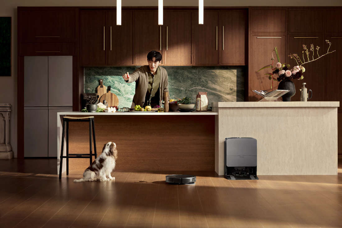 W nowoczesnej kuchni na drewnianej podłodze znajduje się czarny robot sprzątający. Na blacie kuchennym leżą różne produkty spożywcze, a w tle widać drewniane szafki i dekoracje.