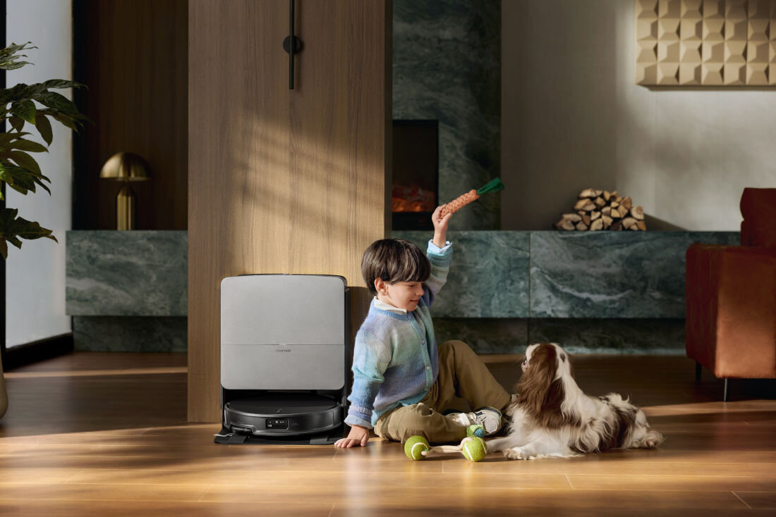 Na drewnianej podłodze w salonie znajduje się robot sprzątający z szarą stacją dokującą. Obok siedzi dziecko z psem, trzymając zabawkę w ręku.