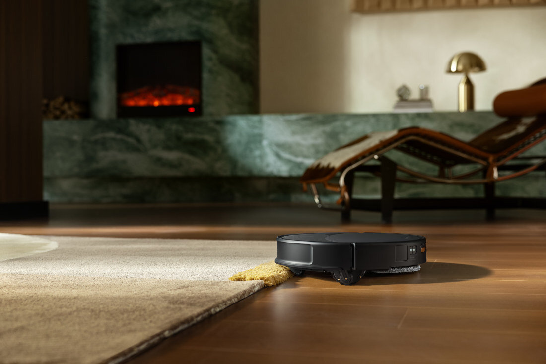 Czarny robot sprzątający porusza się po drewnianej podłodze, częściowo najeżdżając na dywan. W tle widoczny jest nowoczesny salon z kominkiem i leżanką.