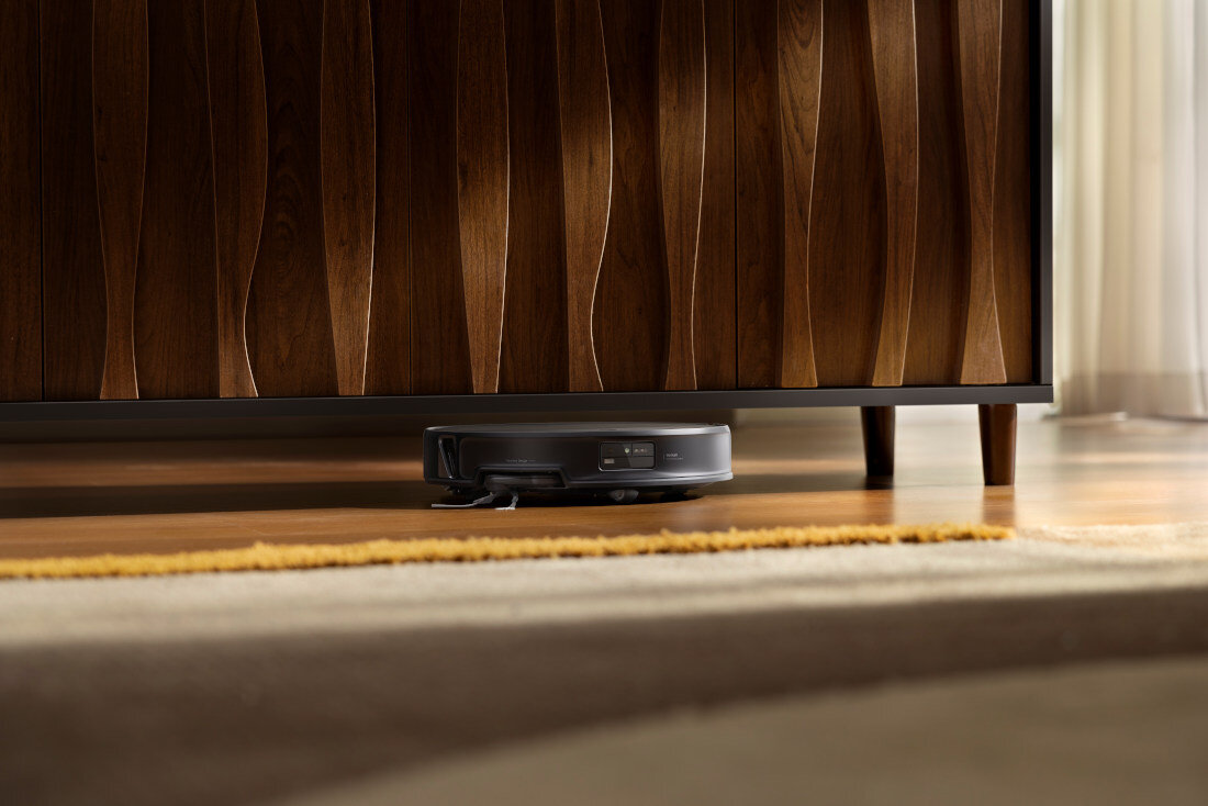 Czarny robot sprzątający znajduje się pod drewnianą szafką z falistym wzorem. Na podłodze leży dywan w kolorze musztardowym.