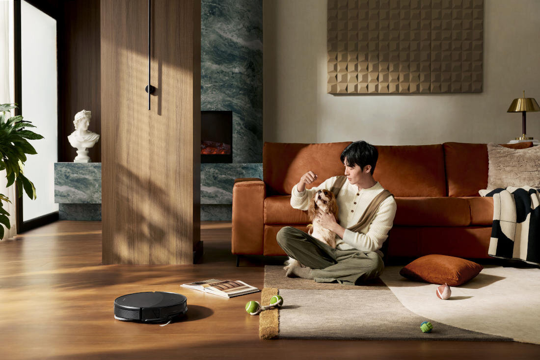 W salonie z brązową sofą i drewnianą podłogą, osoba siedzi na dywanie z psem. Na podłodze znajduje się czarny robot sprzątający oraz kilka zabawek.