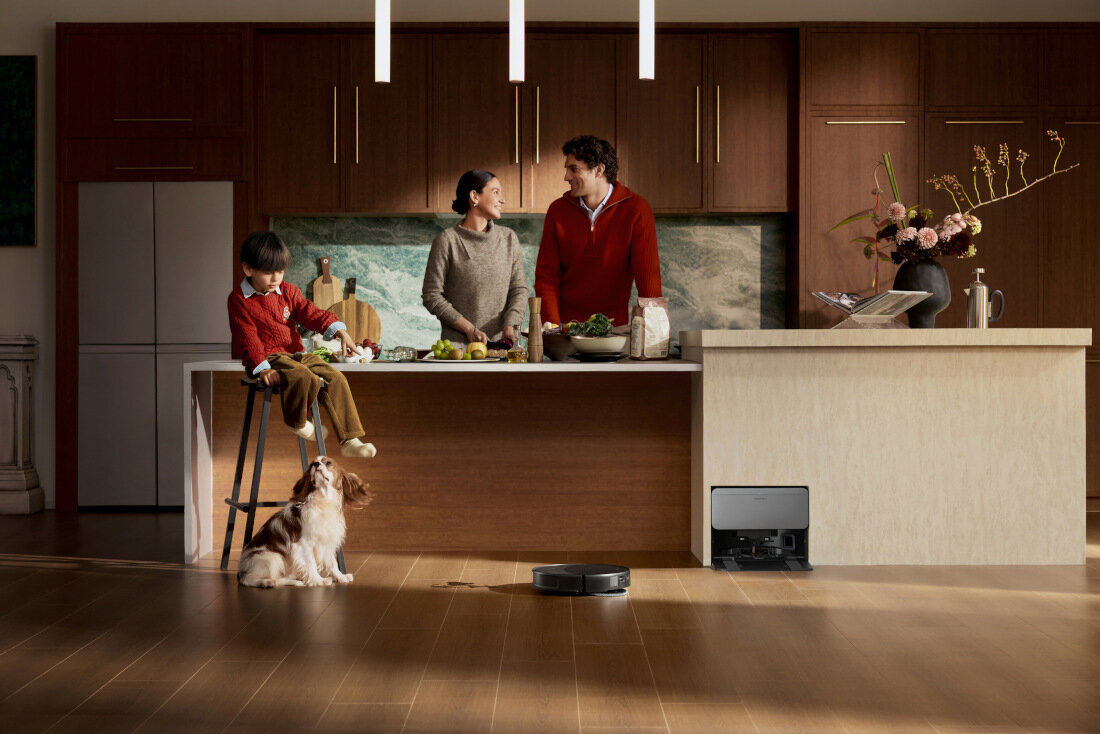 W kuchni znajduje się rodzina przy wyspie kuchennej, na podłodze widoczny jest czarny robot sprzątający. W tle widać drewniane szafki i lodówkę, a na blacie leżą warzywa i kwiaty w wazonie.