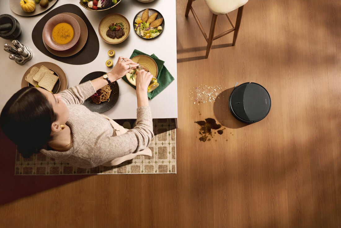 Na drewnianej podłodze znajduje się czarny robot sprzątający obok rozsypanych płatków i rozlanej cieczy. Obok stołu z różnymi potrawami stoi osoba przygotowująca jedzenie.