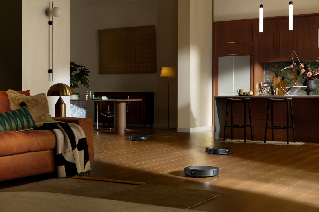 W przestronnym salonie z drewnianą podłogą znajdują się trzy czarne roboty sprzątające. W tle widoczna jest kuchnia z wyspą, krzesłami barowymi i lampami wiszącymi.