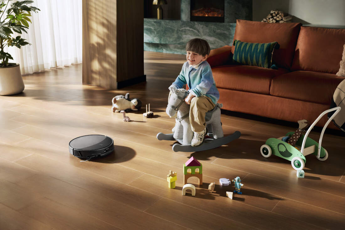Na drewnianej podłodze w salonie znajduje się czarny robot sprzątający oraz rozrzucone zabawki. W tle widać kanapę, roślinę i kominek.